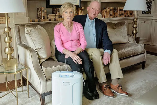 An elderly couple sitting on their couch together with the Inogen At Home system nearby