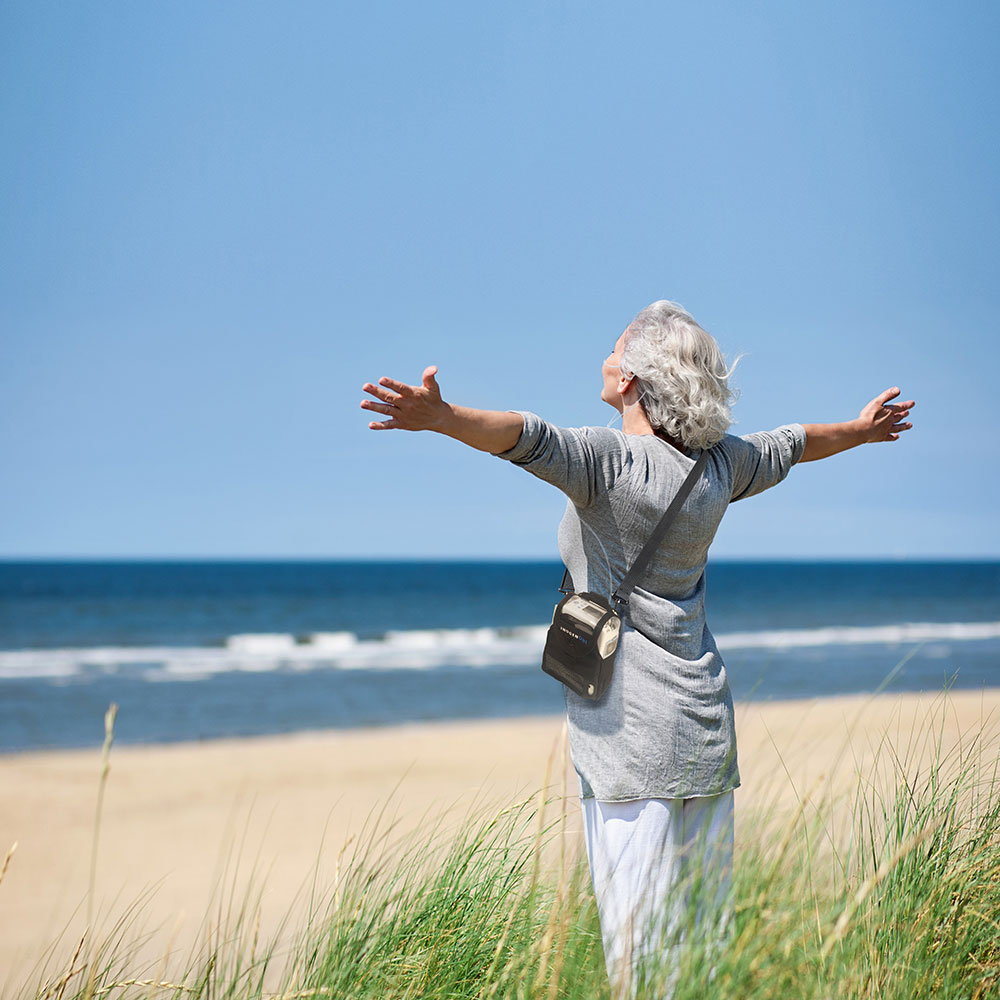 A woman on the beach with arms stretched out wearing an Inogen portable oxygen concentrator